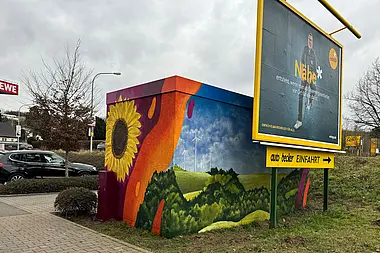 Bunte Malerei auf einer Trafostation zeigt eine Sonnenblume mit Feldern. Davor sieht man ein ENTEGA-Plakat mit einem Mann und dem Wort "Nähe".
