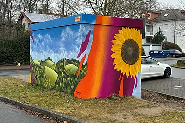 Bunte Trafostation mit Hügellandschaft und Sonnenblume und einem geparkten Auto im Hintergrund.