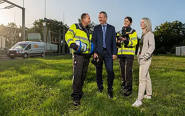 Zwei Mitarbeiterinnen und zwei Mitarbeiter der e-netz Südhessen stehen auf einer Wiese vor der Umspannanlage bei Oder-Roden; im Hintergrund sind ein e-netz-Fahrzeug, Bäume, Büsche und ein strahlend blauer Himmel zu sehen.