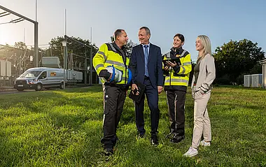 Zwei Mitarbeiterinnen und zwei Mitarbeiter der e-netz Südhessen stehen auf einer Wiese vor der Umspannanlage bei Oder-Roden; im Hintergrund sind ein e-netz-Fahrzeug, Bäume, Büsche und ein strahlend blauer Himmel zu sehen.