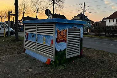 Trafostation mit Graffiti und e-netz Südhessen Schriftzug an einer Straße bei Sonnenuntergang.