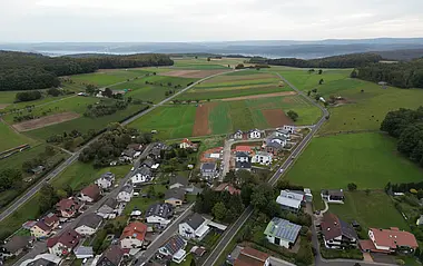 Luftaufnahme zeigt eine Siedlung mit Häusern und Gärten, eingebettet in eine hügelige Landschaft.