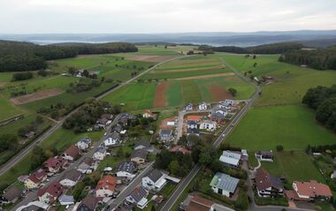 Luftaufnahme zeigt eine Siedlung mit Häusern und Gärten, eingebettet in eine hügelige Landschaft.