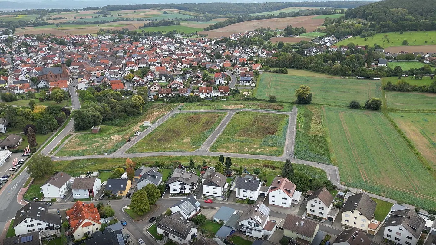 Luftaufnahme zeigt eine Siedlung mit umliegenden Feldern und Grünflächen.