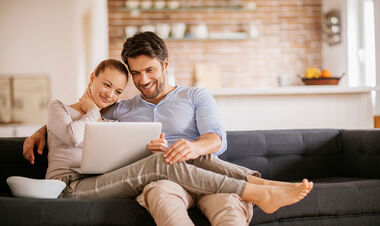 Mann und Frau sitzen auf einer grauen Eckgarnitur und schauen gemeinsam auf den Laptop