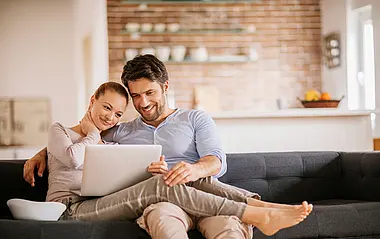 Mann und Frau sitzen auf einer grauen Eckgarnitur und schauen gemeinsam auf den Laptop