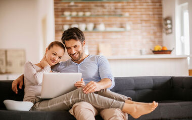 Mann und Frau sitzen auf einer grauen Eckgarnitur und schauen gemeinsam auf den Laptop