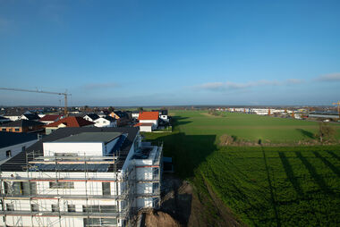 Neubaugebiet mit eingerüstetem Wohnhaus und angrenzenden Feldern an einem sonnigen Tag.
