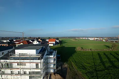 Neubaugebiet mit eingerüstetem Wohnhaus und angrenzenden Feldern an einem sonnigen Tag.