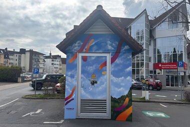 Bunt bemalte Trafostation mit einer Biene und Himmel. Die Station steht auf einem Parkplatz mit Blick auf ein Wohngebäude.