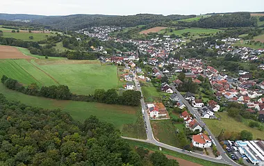 Luftaufnahme zeigt eine ländliche Siedlung mit Feldern und bewaldeten Hügeln.