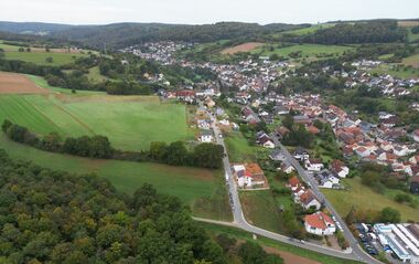 Luftaufnahme zeigt eine ländliche Siedlung mit Feldern und bewaldeten Hügeln.