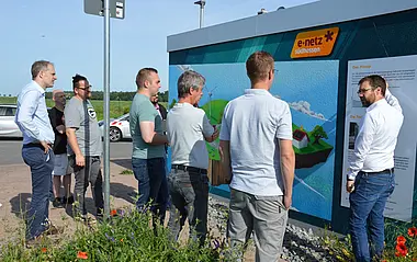 Eine Gruppe von Männern betrachtet eine Informationstafel über erneuerbare Energien im Freien.