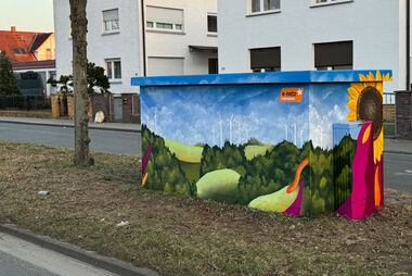 Gestaltete Trafostation mit grüner Hügellandschaft und Baumgruppen und einem blauen Himmel. Im Hintergrund stehen Wohnhäuser.