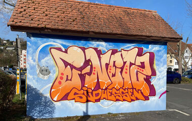 Bunte Trafostation mit einem Graffitischriftzug "e-netz Südhessen", mit Blick auf eine Straße an einem sonnigen Tag.