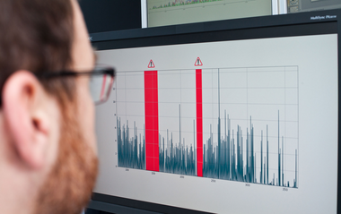 Mitarbeiter sitzt vor einem Bildschirm, auf dem ein technischer Graph mit roten Warnhinweisen angezeigt wird.