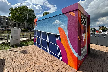 Bunt gestaltete Trafostation mit Blick auf eine Stadtlandschaft.