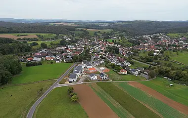 Luftaufnahme zeigt landwirtschaftliche Flächen und eine kleine Siedlung im Grünen.