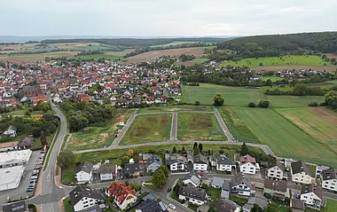 Luftaufnahme zeigt eine Wohngegend mit Gärten und Feldern vor dem Hintergrund bewaldeter Hügel.