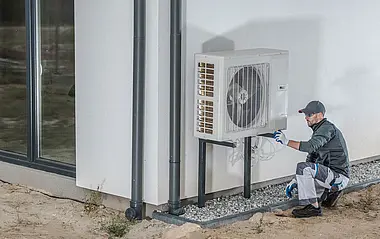 Ein Techniker installiert eine Wärmepumpe an einer weißen Fassade.