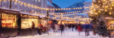Verschneiter Weihnachtsmarkt bei Abenddämmerung mit Besuchern und beleuchtetem Weihnachtsbaum