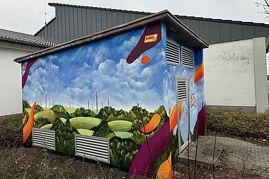 Gestaltete Trafostation mit Windkraftanlagen in einer hügeligen Landschaft bei bedecktem Himmel und e-netz Südhessen-Logo.