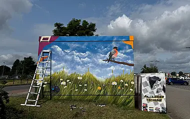 Künstlerisch gestaltete Trafostation auf einem Parkplatz, umgeben von einer blühenden Wiese. Ein Vogel sitzt in der Nähe, darüber ein bewölkter Himmel.
