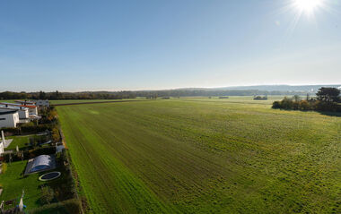 Grünes Feld mit Gebäuden am Rand und Hügeln im Hintergrund.