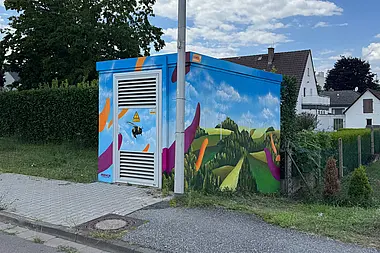 Bunt bemalte Trafostation mit einer Biene, umgeben von Feldern und Bäumen. Im Hintergrund stehen Wohnhäuser, die von Büschen eingerahmt werden.