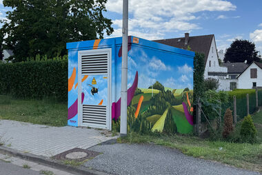 Bunt bemalte Trafostation mit einer Biene, umgeben von Feldern und Bäumen. Im Hintergrund stehen Wohnhäuser, die von Büschen eingerahmt werden.