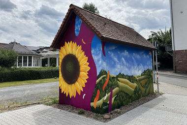 Gestaltete Trafostation mit Sonnenblume, Feldern und bewölkten Himmel mit Blick auf Wohnhäuser im Hintergrund.