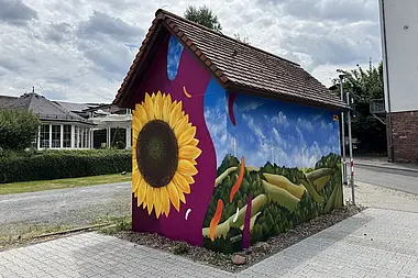 Gestaltete Trafostation mit Sonnenblume, Feldern und bewölkten Himmel mit Blick auf Wohnhäuser im Hintergrund.
