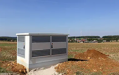 Verteilungsstation auf Baustelle, umgeben von Erde, mit Blick auf Windkraftanlagen.