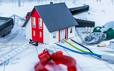 Spielzeughaus mit grauem Dach steht auf einem Bauplan und ist umringt von verschiedenen Werkzeugen